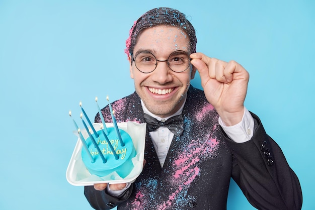 La foto de un hombre adulto guapo celebra el cumpleaños contento de recibir felicitaciones lleva gafas redondas traje formal sonríe felizmente posa con un delicioso pastel contra el fondo azul Concepto de vacaciones