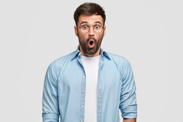 Foto de hombre sin afeitar aterrorizado mira con ojos llenos de incredulidad, siendo sorprendido por una revelación curiosa, viste camisa azul, aislado sobre una pared blanca. Personas, reacción, expresiones faciales.