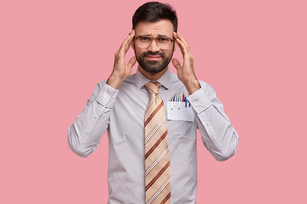 Foto de hombre sin afeitar descontento que sufre de dolor de cabeza, mantiene las manos en las sienes, tiene expresión de tristeza, cansado después del trabajo en la oficina