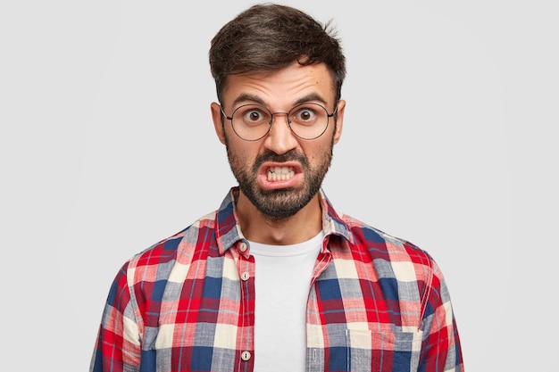 Foto de hombre sin afeitar irritado que se ve enojado, aprieta los dientes y levanta las cejas, está molesto con muchas obligaciones en el trabajo, vestido con camisa a cuadros, se encuentra contra la pared blanca.