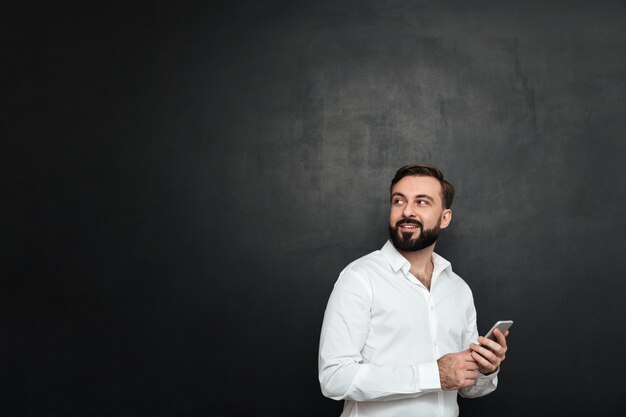 Foto de hombre alegre con camisa blanca mirando hacia atrás mientras chatea o usa la conexión inalámbrica en el teléfono móvil sobre gris oscuro