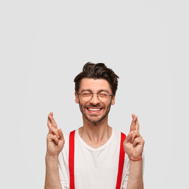 Foto de hombre alegre guapo sin afeitar con corte de pelo moderno cruza los dedos, cree en algo fino, mantiene los ojos cerrados, vestido elegantemente, aislado sobre una pared blanca. Concepto de personas y deseos