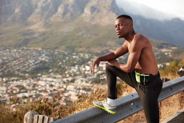 Foto de un hombre atleta con piel oscura y saludable, descansa después de los ejercicios físicos, mantiene las piernas levantadas en la señal de tráfico, tiene una expresión pensativa, posa en las montañas y disfruta del deporte al aire libre. Concepto de jogging