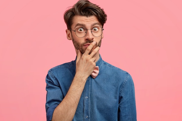 Foto de hombre barbudo caucásico asustado sorprendido mantiene la mano en la boca, mira con ojos llenos de conmoción, viste elegante camisa de mezclilla