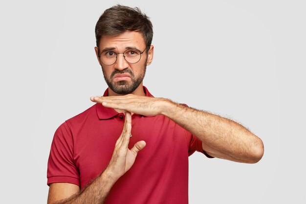 La foto de un hombre barbudo disgustado mantiene las manos perpendicularmente, tiene una expresión incierta, trata de explicar la ruta o la dirección, usa gafas redondas y una camiseta roja, aislada sobre una pared blanca