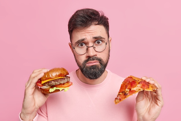 La foto del hombre barbudo disgustado no puede negarse a comer comida rápida tiene una deliciosa hamburguesa y una rebanada de pizza sabrosa se ve triste, tiene una nutrición poco saludable