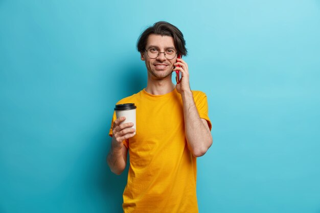 Foto de hombre europeo adulto guapo tiene conversación telefónica a través de un teléfono inteligente bebe café para llevar