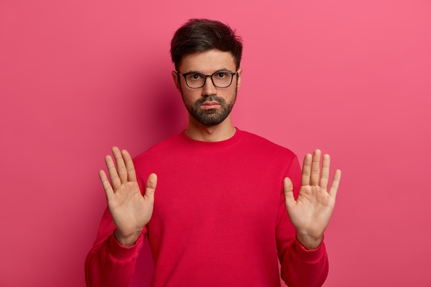 La foto de un hombre europeo con una barba densa hace un gesto de alto, sostiene un cartel, tira de las palmas de las manos, mira con seriedad, tiene una expresión decidida, pide calmarse, usa un suéter rojo, gafas.