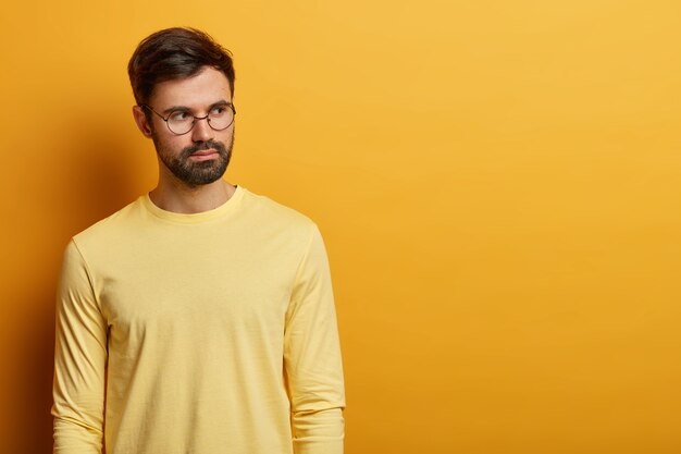 foto de un hombre europeo barbudo pensativo reflexiona sobre la decisión, usa gafas redondas y un suéter amarillo informal, copia espacio para contenido publicitario, piensa en la idea, tiene expresión facial tranquila.