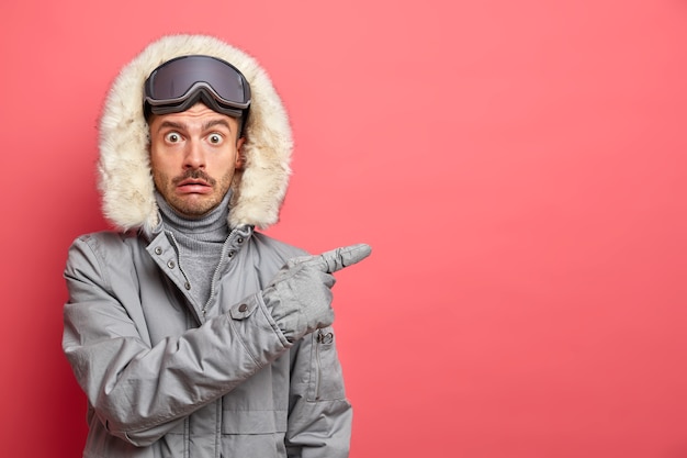La foto del hombre europeo emocional conmocionado vestido con ropa de invierno usa gafas de esquí y apunta hacia el espacio en blanco da la dirección a la derecha.