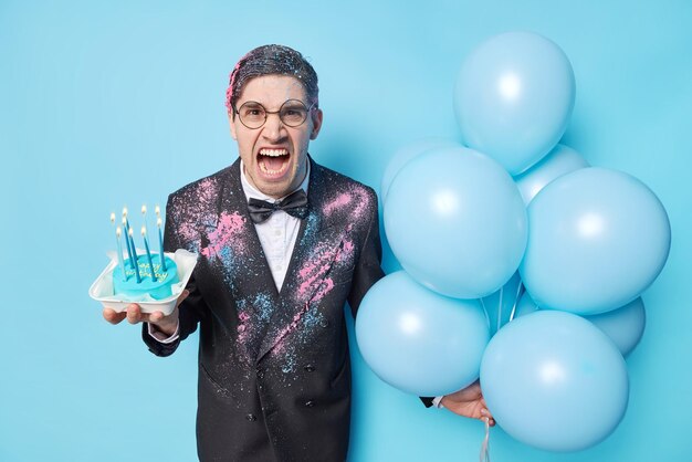 La foto de un hombre europeo irritado exclama en voz alta con molestia celebra el cumpleaños sostiene un pastel con velas encendidas y globos inflados vestidos con traje festivo aislado sobre fondo azul