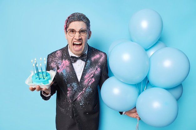 La foto de un hombre europeo irritado exclama en voz alta con molestia celebra el cumpleaños sostiene un pastel con velas encendidas y globos inflados vestidos con traje festivo aislado sobre fondo azul