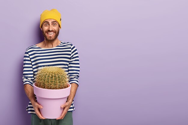 Foto de hombre feliz con una sonrisa agradable, sostiene una maceta de cactus espinosos, está de buen humor, vestido con un suéter a rayas, posa sobre un fondo púrpura con espacio libre