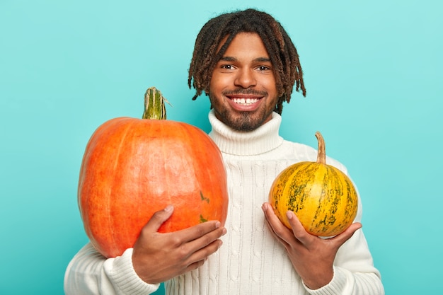 Foto de hombre guapo alegre hipster con rastas, sonrisa con dientes, sostiene dos calabazas de diferentes tamaños, viste un suéter blanco cálido, aislado sobre una pared azul