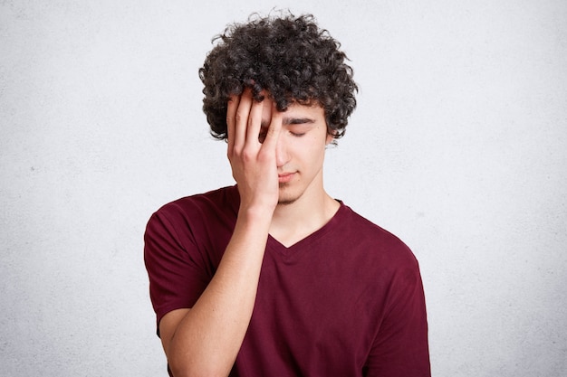 Foto de hombre guapo cansado con exceso de trabajo con cabello oscuro y rizado, cubre la cara con la mano, se ve frustrado. Adolescente mantiene los ojos cerrados