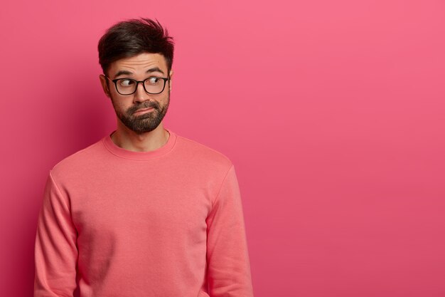 Foto de un hombre guapo y pensativo con una espesa barba oscura concentrada a un lado en el espacio vacío de la pared rosa, nota una escena interesante, tiene una expresión de sorpresa, usa lentes transparentes y un jersey.