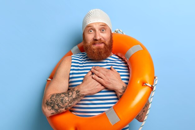 Foto de hombre impresionado conmovido que mantiene las manos cerca del corazón, promete algo a su esposa, usa gorra de goma y chaleco de marinero a rayas