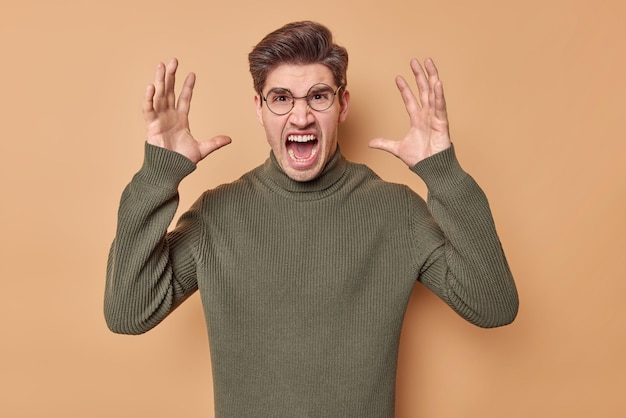 La foto de un hombre indignado grita de molestia grita en voz alta mantiene las palmas levantadas expresa emociones negativas usa anteojos redondos y suéter aislado sobre fondo beige Modelo masculino furioso