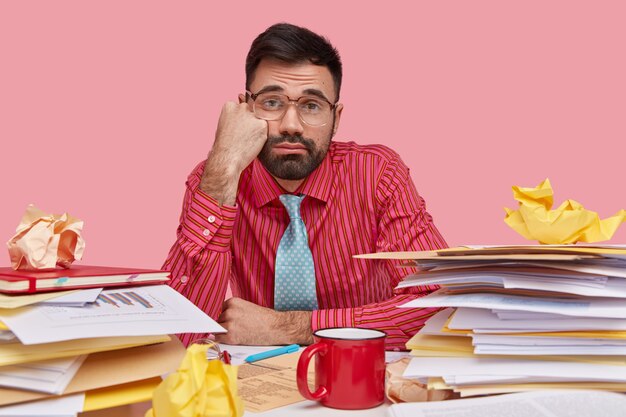 La foto del hombre insatisfecho con sueño tiene la mano en la mejilla, se ve con expresión triste, usa camisa rosa, gafas grandes, bebe café o té, tiene muchos papeles en la mesa