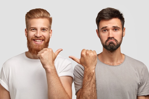 Foto de hombre de jengibre joven alegre y punto de chico caucásico sin afeitar disgustado con los pulgares el uno al otro, trabajar juntos como equipo, aislado sobre pared blanca. Concepto de amistad y emociones