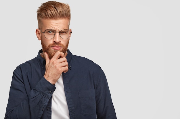 Foto de hombre jengibre serio brutal con barba espesa, sostiene la barbilla, levanta las cejas, está sumido en pensamientos, contempla algo, aislado sobre una pared blanca