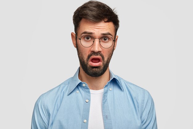 Foto de hombre joven con barba oscura que hace la cara aburrida, escucha algo con falta de interés, tiene expresión de descontento, vestido con camisa azul, abre la boca con disgusto, aislado sobre una pared blanca