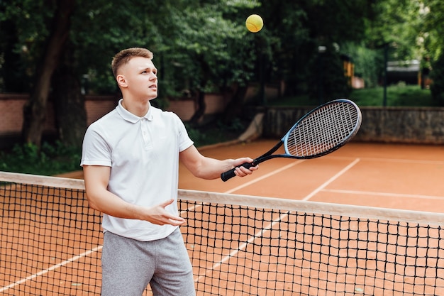 Foto de hombre joven feliz en camisa de polo con raqueta de tenis y sonriendo mientras está de pie en la cancha de tenis.