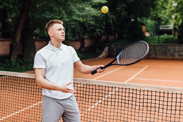 Foto de hombre joven feliz en camisa de polo con raqueta de tenis y sonriendo mientras está de pie en la cancha de tenis.