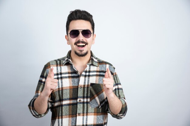 Foto de hombre joven con gafas negras de pie sobre una pared blanca.