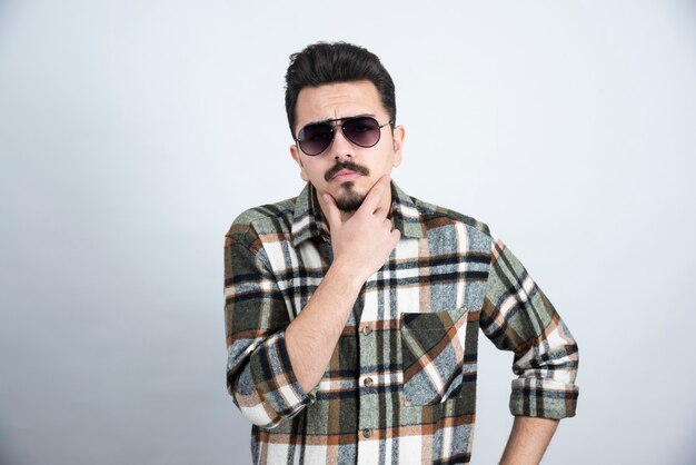 Foto de hombre joven con gafas negras de pie sobre una pared blanca.