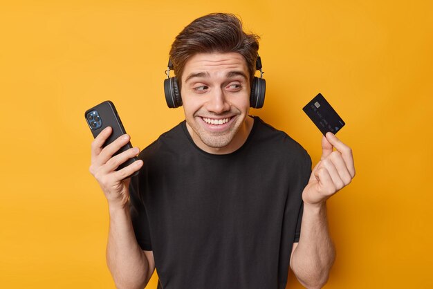 Foto de hombre moreno feliz posa con tarjeta de crédito y pedidos de teléfonos inteligentes en la aplicación paga en línea escucha música a través de auriculares inalámbricos vestidos con una camiseta negra informal aislada sobre fondo amarillo