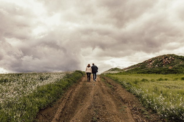 Foto de un hombre y una mujer caminando por un sendero en un valle con flores bajo un cielo brumoso