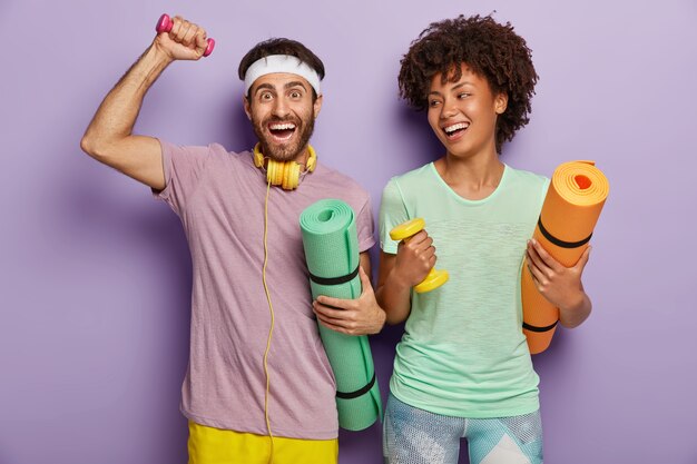 Foto de hombre y mujer felices que trabajan en bíceps con pesas, cargan karemats, tienen expresiones alegres, disfrutan entrenar juntos, se visten con ropa casual, se motivan para un estilo de vida saludable y el deporte