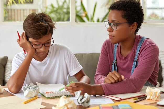 Foto de hombre y mujer de raza mixta sentarse juntos en el lugar de trabajo, discutir ideas para el proyecto, usar gafas. La mujer negra con piercing explica algo al hermano, indica en escritos en el diario.