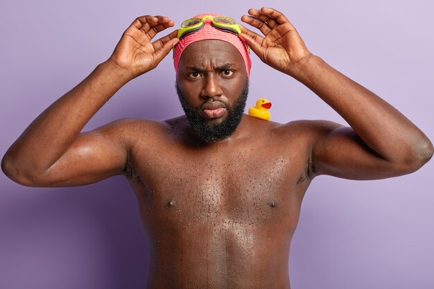 La foto de un hombre negro enojado ajusta las gafas, se ve triste, asiste a la sección de deportes, nada muy bien, tiene un patito de goma en el hombro, sale del agua tiene la piel húmeda, se ve insatisfecho