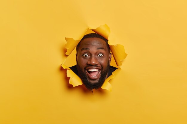 Foto de hombre negro feliz con expresión facial complacida, cerdas oscuras, se divierte en el interior, mantiene la cabeza en el agujero de papel rasgado, se ríe y mira a la cámara, aislada sobre fondo amarillo.