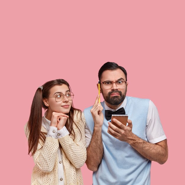 Foto de un hombre pensativo con cerdas gruesas, sostiene un bloc de notas y un lápiz, piensa en ideas para escribir un ensayo, su novia geek intenta ayudar, se paran juntos, trabajan juntos, aislados en rosa