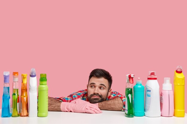 Foto de hombre pensativo disgustado cansado se inclina sobre la mesa, concentrado en botellas de agente de limpieza, viste camisa a cuadros