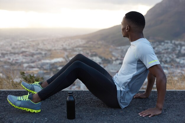 Foto de hombre relajado con cabello corto, piel oscura, sentado en la carretera con una botella de agua fría, viste chándal, concentrado a un lado, disfruta del tiempo libre, vista a la montaña, descansa después del entrenamiento. Fitness, ejercicio