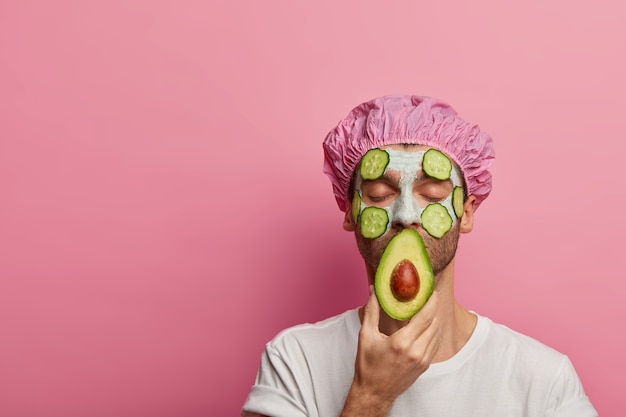 Foto de hombre tranquilo y satisfecho disfruta de tratamientos de belleza en el salón de spa, sostiene aguacate, usa máscara vegetal, tiene procedimientos de cosmetología