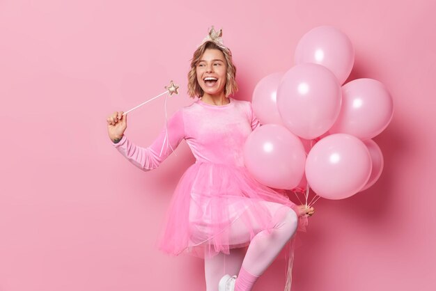 Una foto horizontal de una alegre joven rubia vestida con ropa festiva sostiene una varita mágica y un montón de globos celebra el cumpleaños feliz de conocer a los invitados en poses de fiesta con fondo rosa