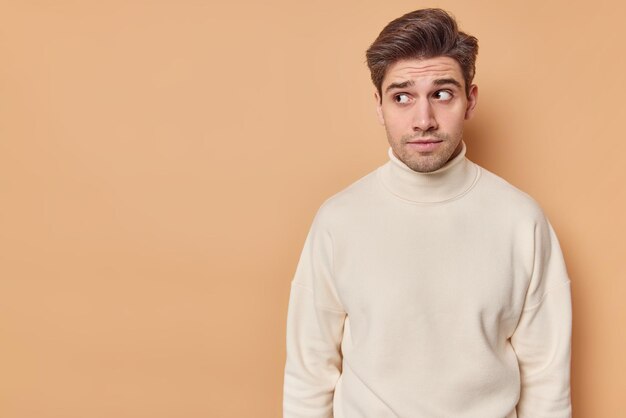 La foto horizontal de un apuesto hombre de cabello oscuro mira con expresión curiosa algo interesante que se siente sospechoso, usa un cuello de tortuga casual aislado sobre una pared beige con espacio para copiar texto