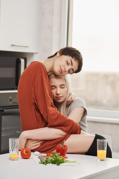 Foto horizontal de dos mujeres abrazándose en la cocina con los ojos cerrados y una sonrisa sensual, sentado en la mesa. Las encantadoras chicas enamoradas finalmente se abrieron sobre sus sentimientos entre ellas.