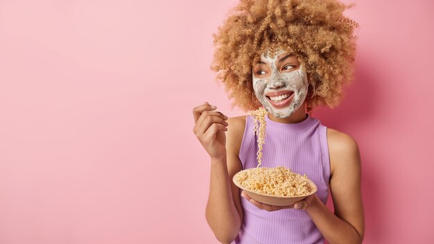 Una foto horizontal de una joven alegre que come pasta del tazón se ve felizmente lejos, se somete a procedimientos de belleza y aplica una máscara nutritiva de arcilla en la cara aislada en un espacio vacío de fondo rosa para su texto