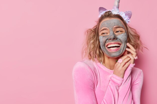 Una foto horizontal de una joven europea feliz con una mascarilla facial nutritiva que se somete a procedimientos de cuidado de la piel mira hacia otro lado y sonríe ampliamente posa contra un fondo rosa con espacio para copiar para su promoción