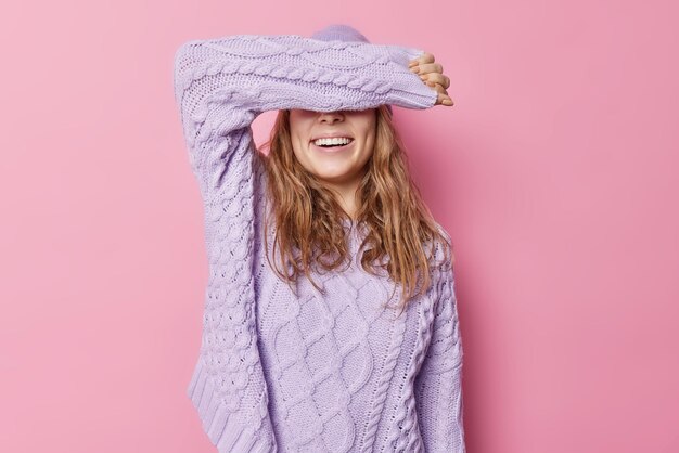 Foto gratuita la foto horizontal de una joven feliz cubre los ojos con el brazo y usa un suéter de punto suelto que trata de ocultar la cara se mantiene positiva aislada sobre un fondo rosado una modelo femenina alegre espera una sorpresa