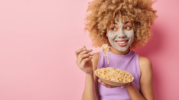 Una foto horizontal de una joven guapa con el pelo rizado aplica una máscara de belleza en la cara y se somete a tratamientos para el cuidado de la piel come deliciosos espaguetis con una camiseta informal aislada en un fondo rosa