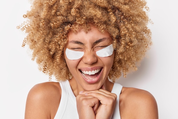 La foto horizontal de una joven de pelo rizado muy contenta mantiene las manos debajo de la barbilla y aplica parches blancos de belleza debajo de los ojos para quitar las bolsas o las poses de arrugas en el interior. Concepto de rutina de cuidado de la piel por la mañana