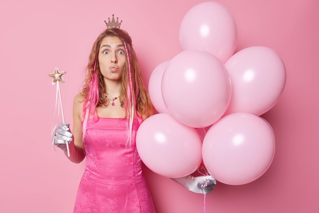 Una foto horizontal de una joven sorprendida usa un vestido festivo de la corona y los guantes mantienen los labios doblados celebra una ocasión especial con un montón de globos de helio y una varita mágica aislada sobre un fondo rosa