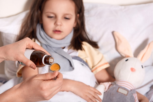 Foto horizontal de mano desconocida vertiendo líquido en la cuchara de una pequeña botella con jarabe, persona que cuida al niño acostado en la cama con gripe
